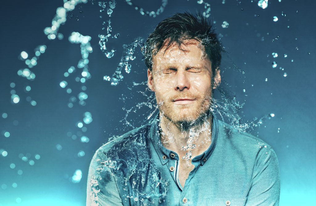 Man with eyes closed as water splashes around his face, wearing a teal shirt against a blue gradient background