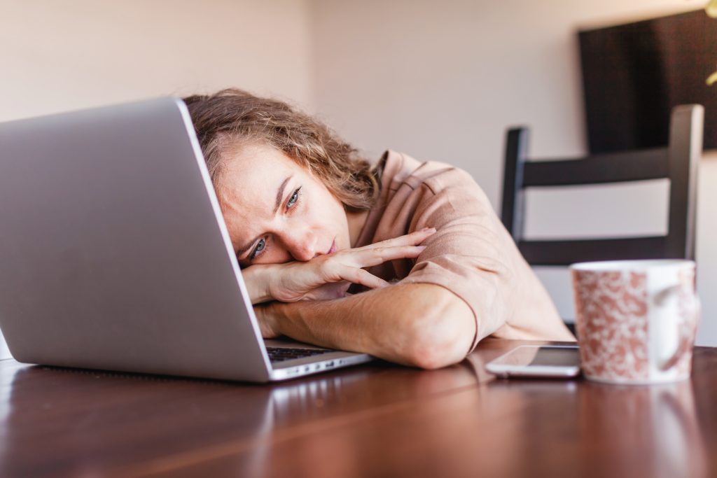 Woman resting her head on a laptop at a wooden desk, looking fatigued.